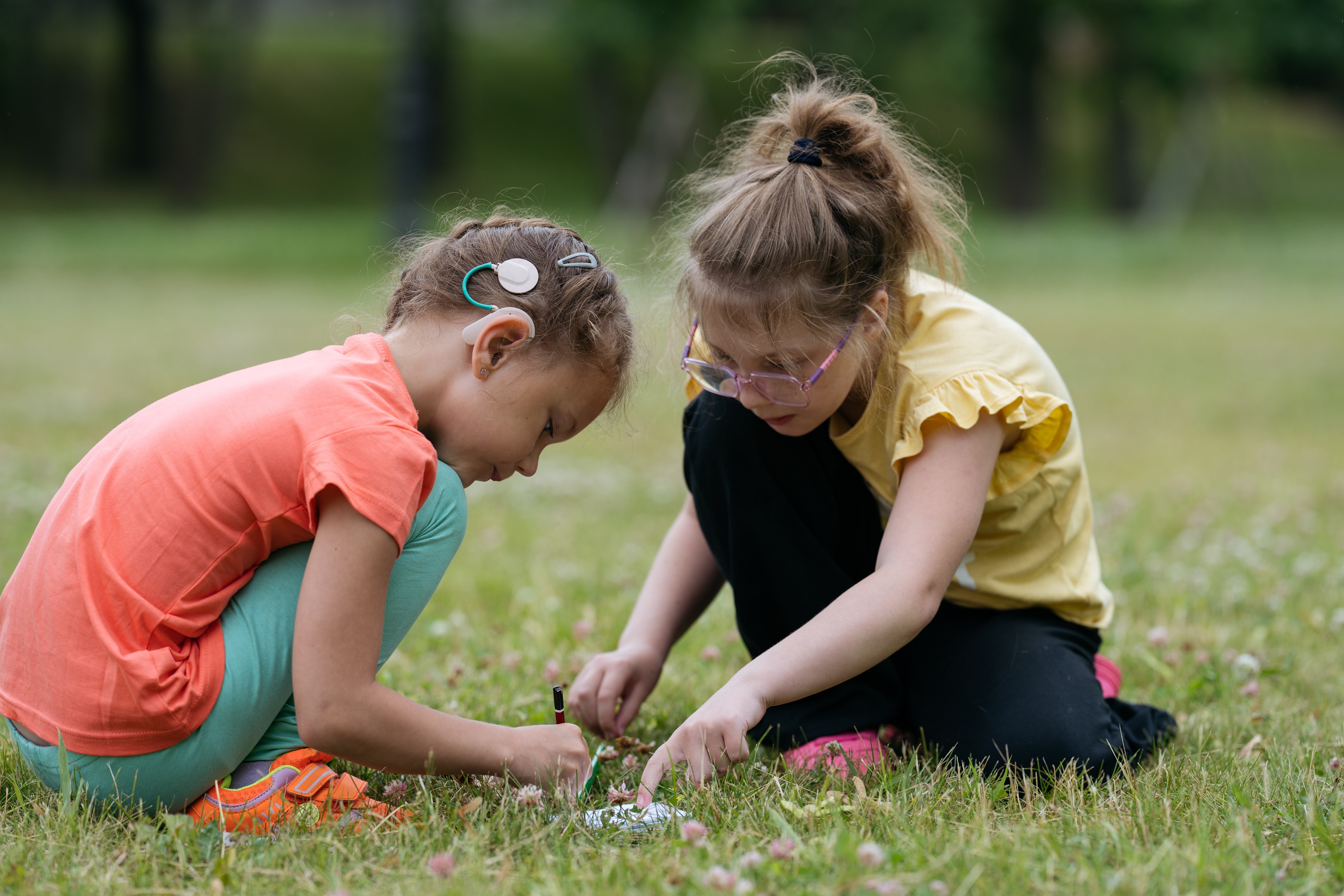 Girl with cochlear implant plays in grass with friend wearing glasses.