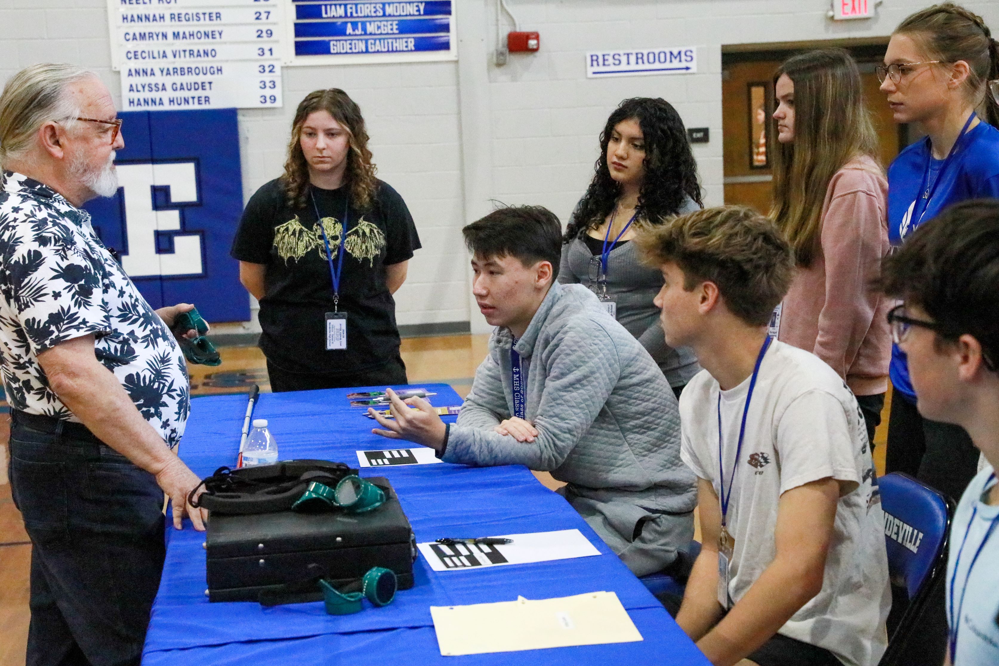 Michael Norman stands behind a table with vision googles and signing guides while several high school students sit in front of the table.