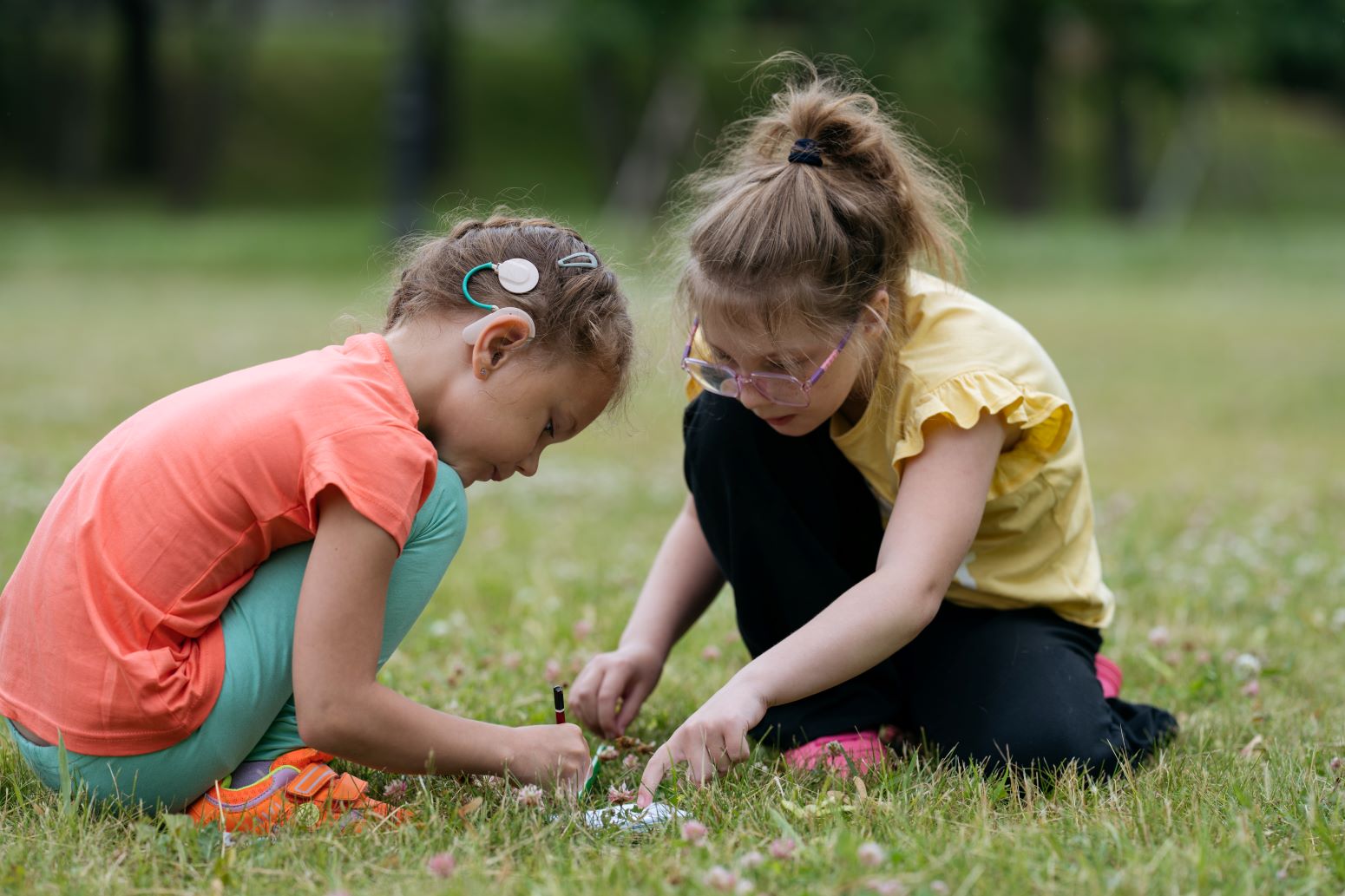 Girl with cochlear implant plays in grass with another girl wearing glasses.
