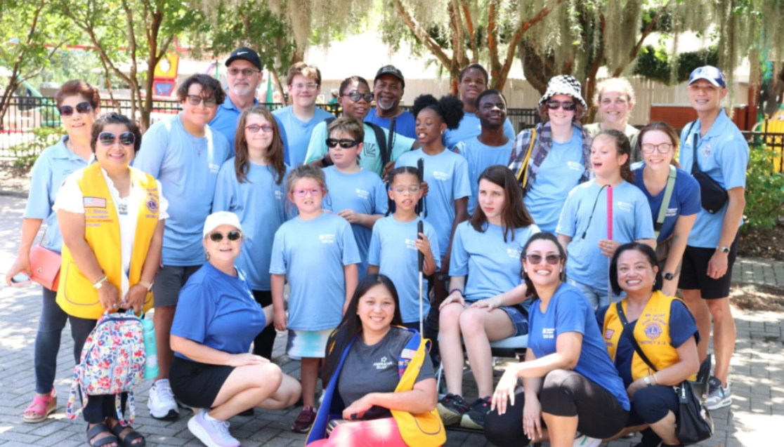 Summer campers and staff wear blue tshirts in a group. Some campers have white canes and wear glasses and sunglasses.