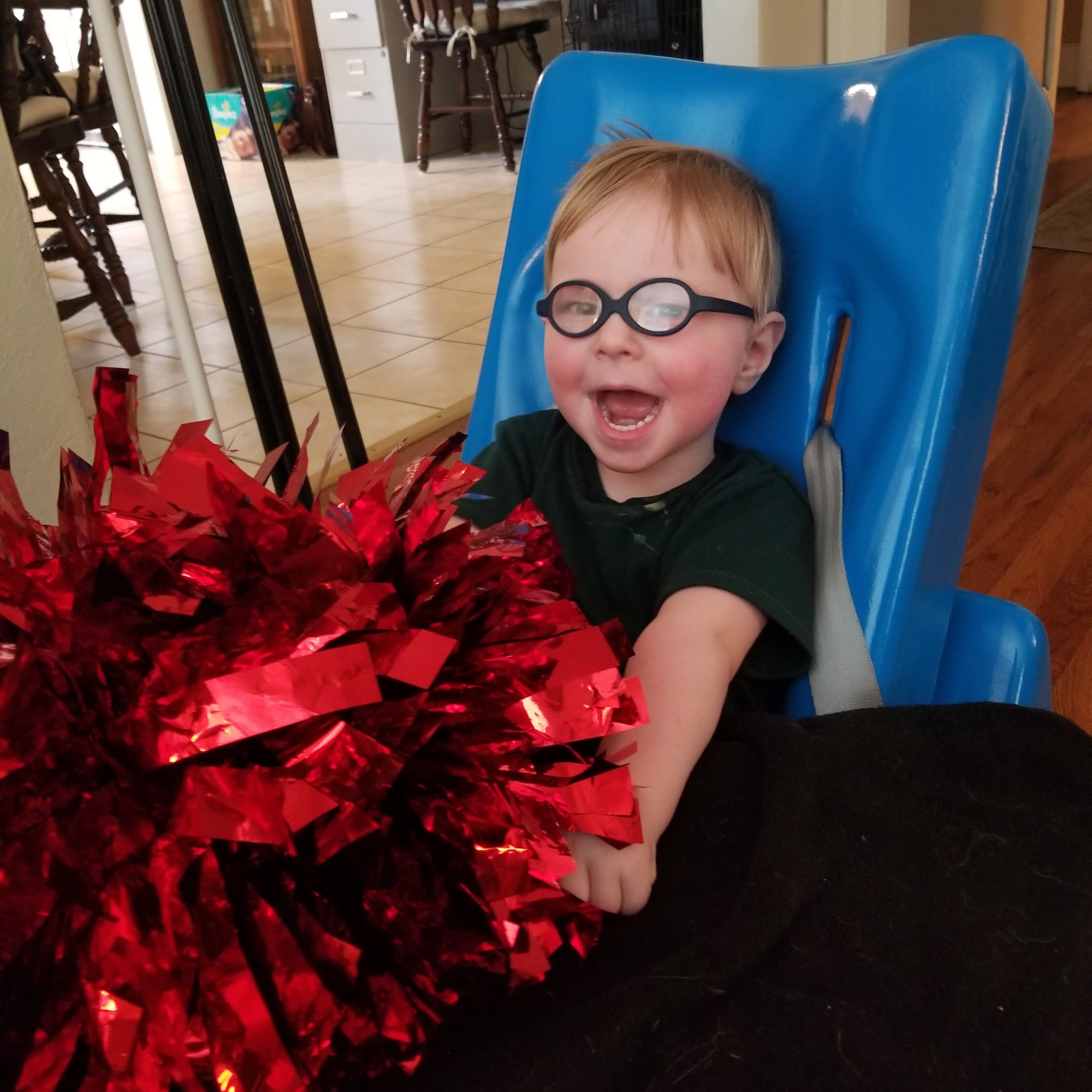 Toddler wearing glasses smiles excitedly as he plays with flashy red pompom.
