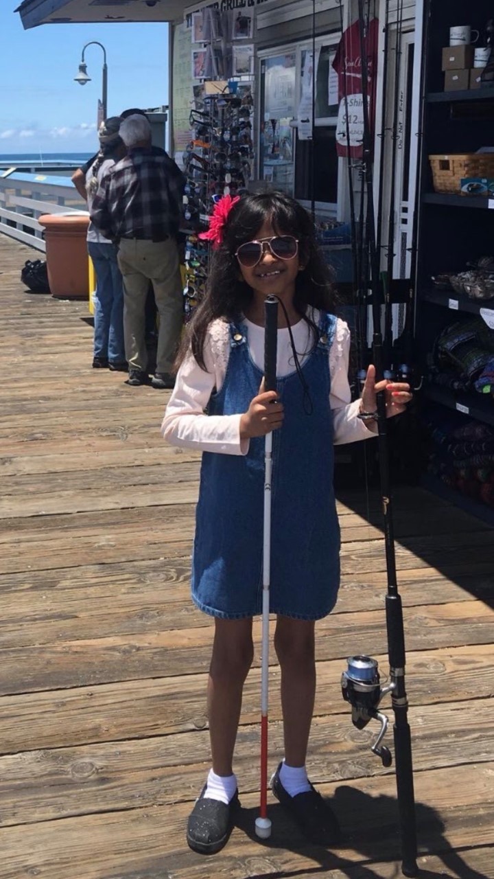 Girl stands on pier holding white cane and a fishing pole while wearing sunglasses.