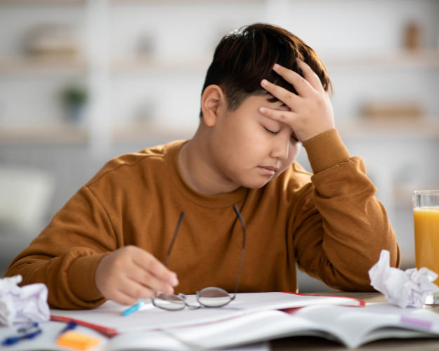 Boy working on schoolwork at home sits with his head in his hand, eyes closed, while holding his eyeglasses.