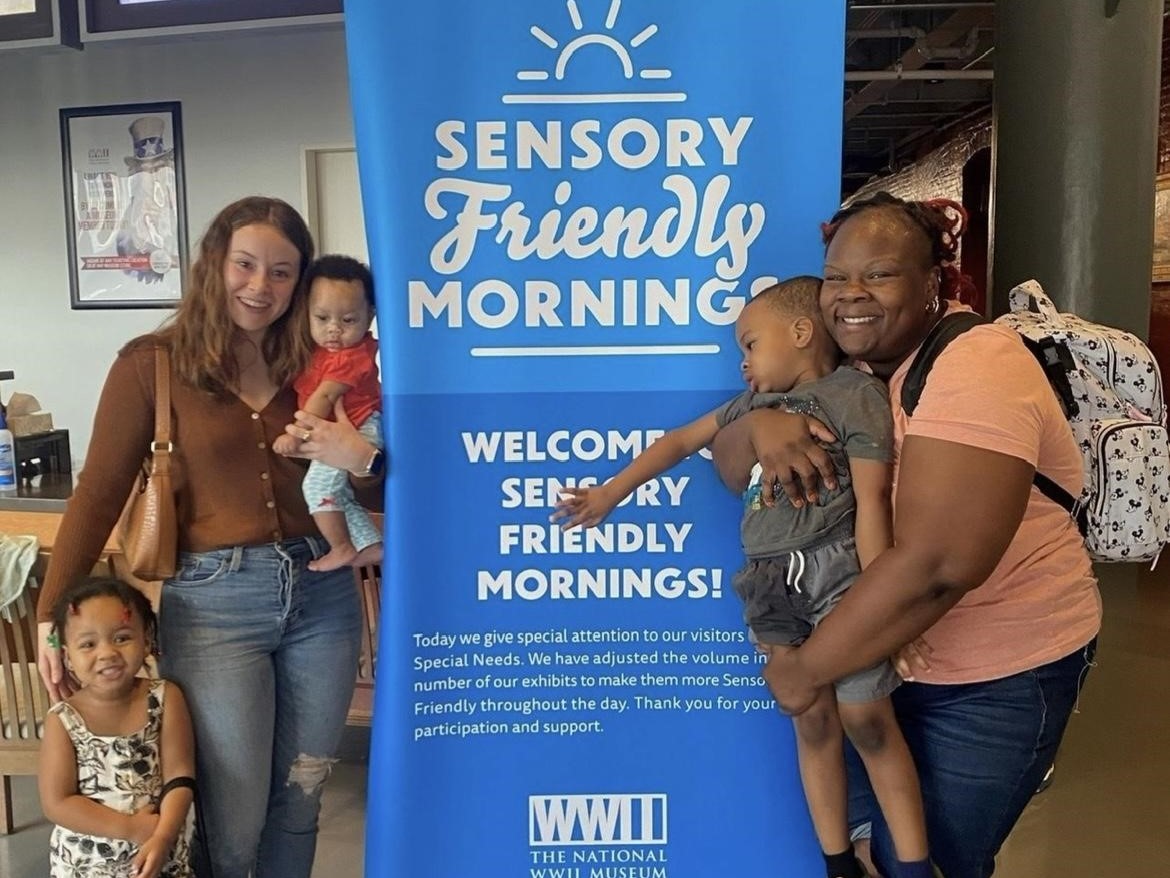 Scholar poses with family of mother and three kids in front of the sesnory-friendly WW2 museum banner.