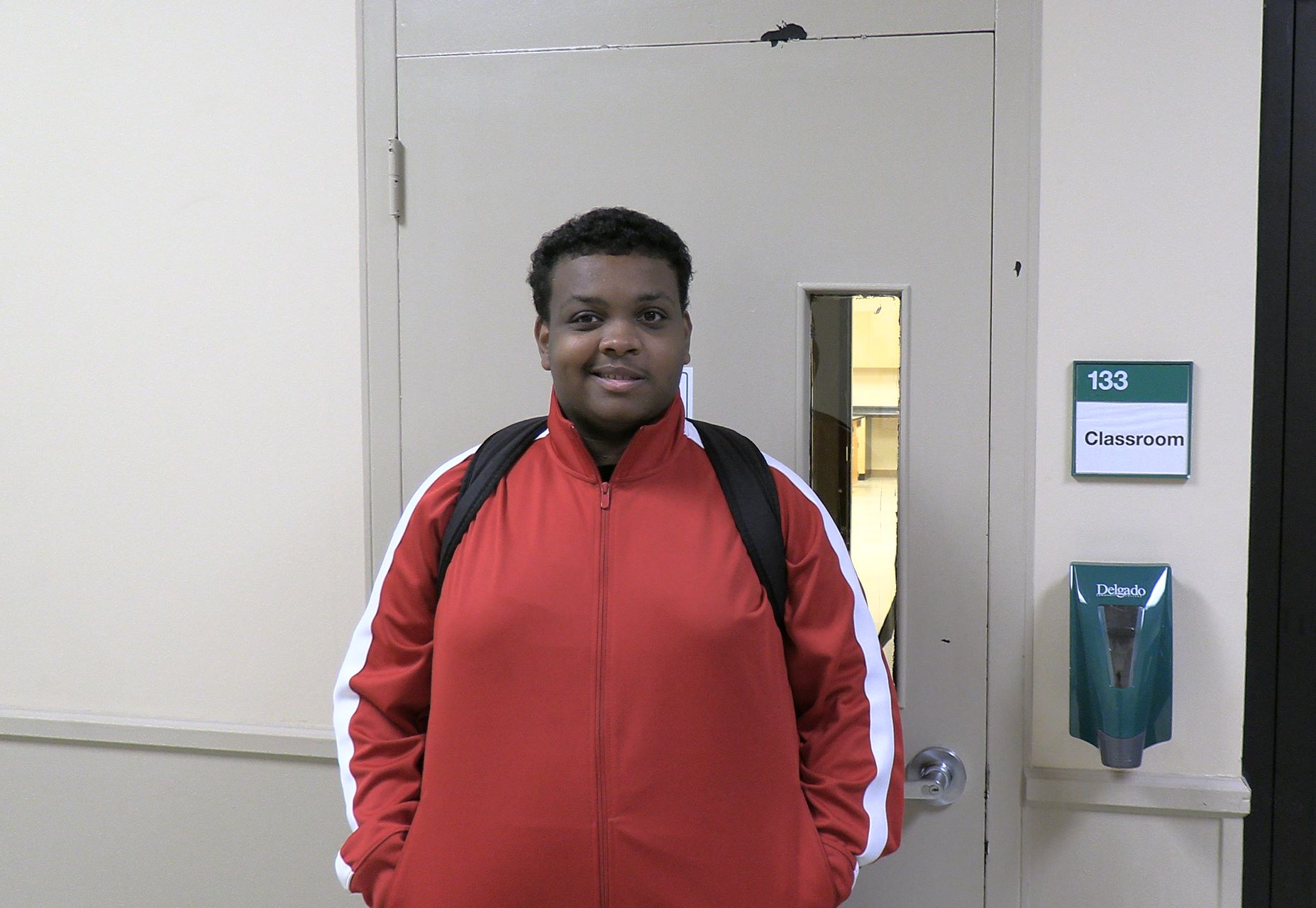 PAYCheck student wears backpack while he stands smiling outside a Delgado classroom.