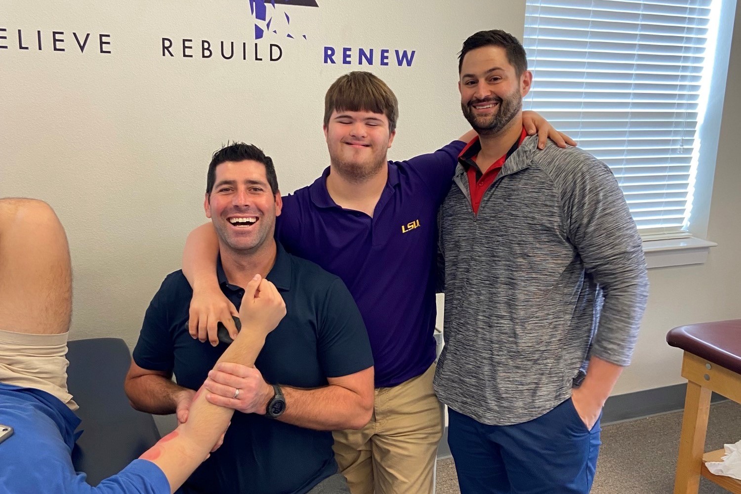 Young man with Down syndrome smiles as he puts his arms around coworkers at a physical therapy clinic. One PT therapist is massaging client's arm. 